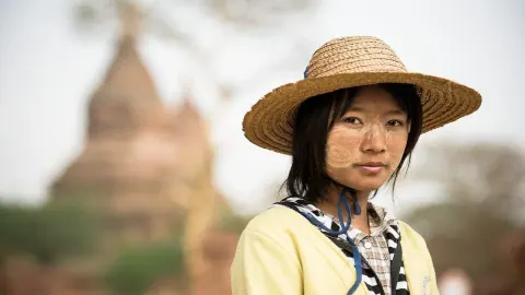 Young Burmese woman wearing thanaka and a straw hat in front of a temple in Bagan, Myanmar